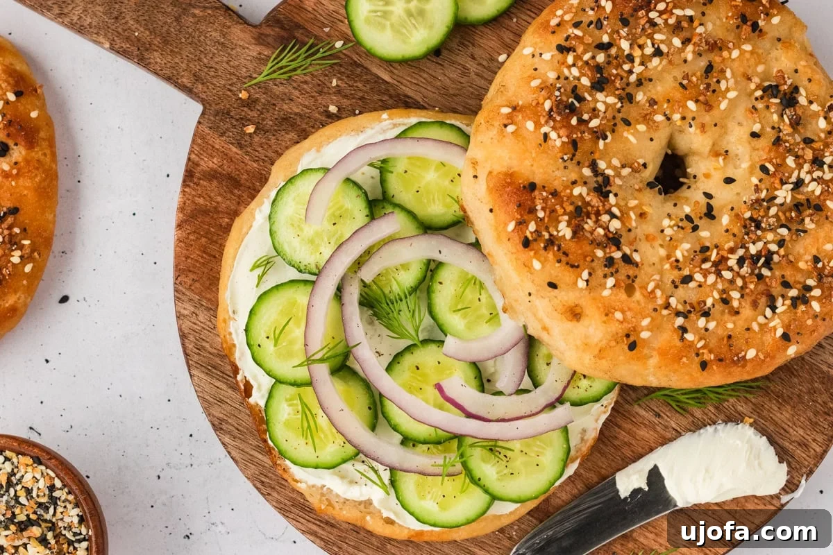 A gluten-free everything bagel sandwich on a wooden board, filled with cream cheese, cucumber slices, red onion, and fresh dill, with a knife and extra cream cheese nearby.