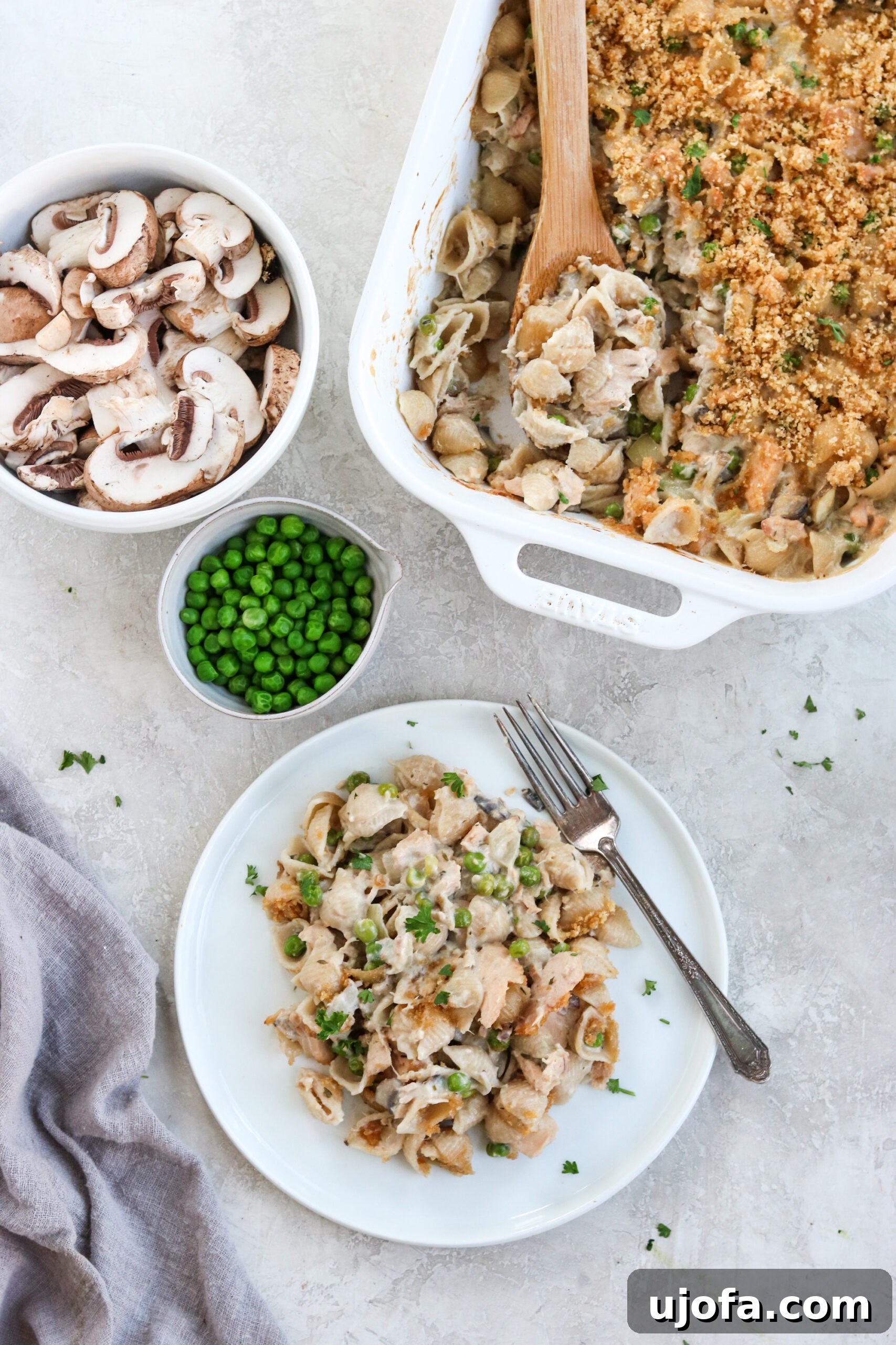 Tuna noodle casserole served on a white plate with the white casserole dish and a bowl of mushrooms and peas on the side