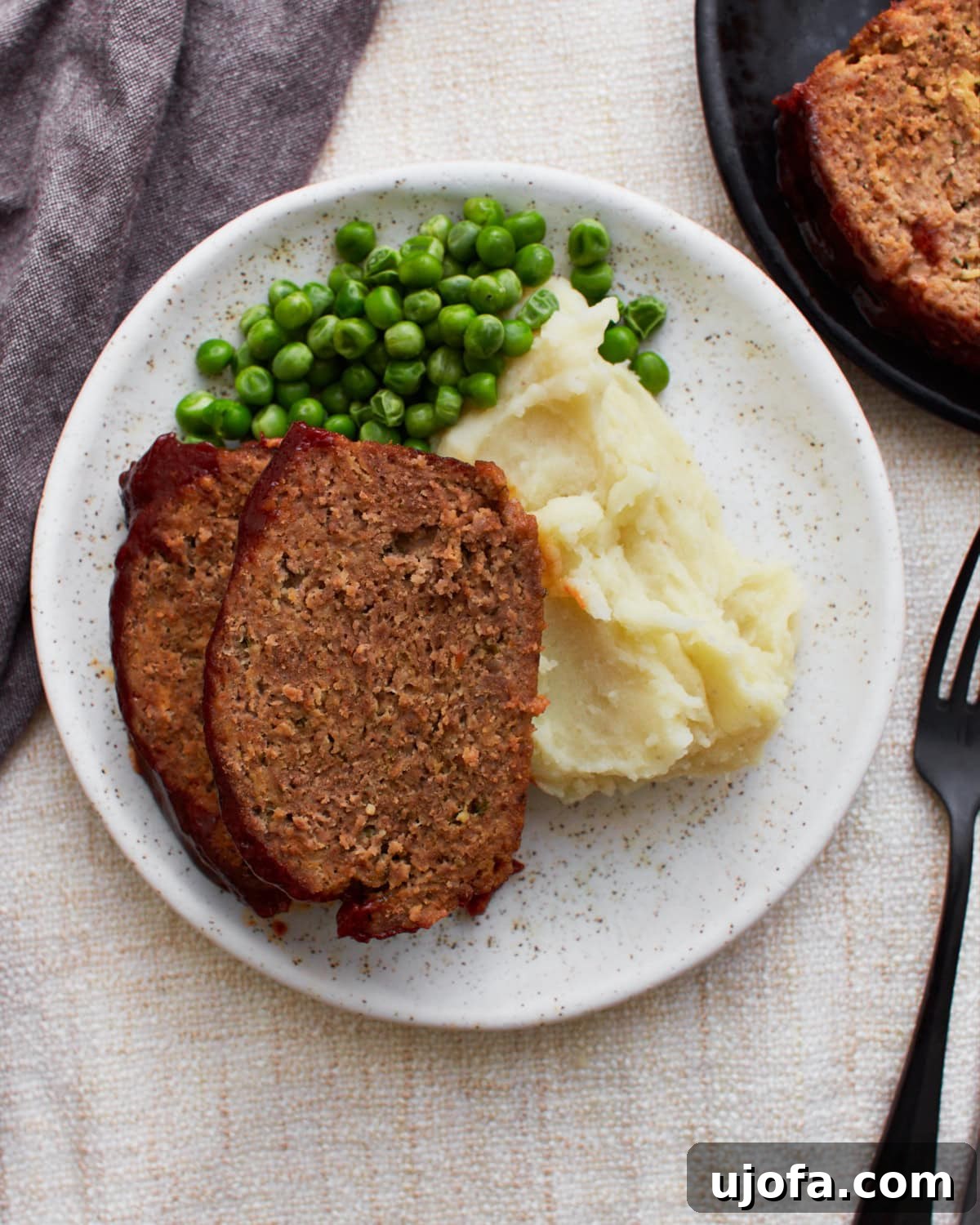 Two slices of glazed gluten-free meatloaf served with creamy mashed potatoes and green peas on a white plate, showcasing a comforting homemade meal.