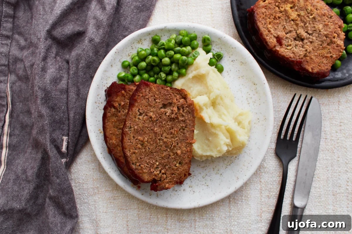 A white plate with two slices of glazed gluten-free meatloaf, creamy mashed potatoes, and green peas, accompanied by a fork and knife, with a cloth napkin and another plate in the background.