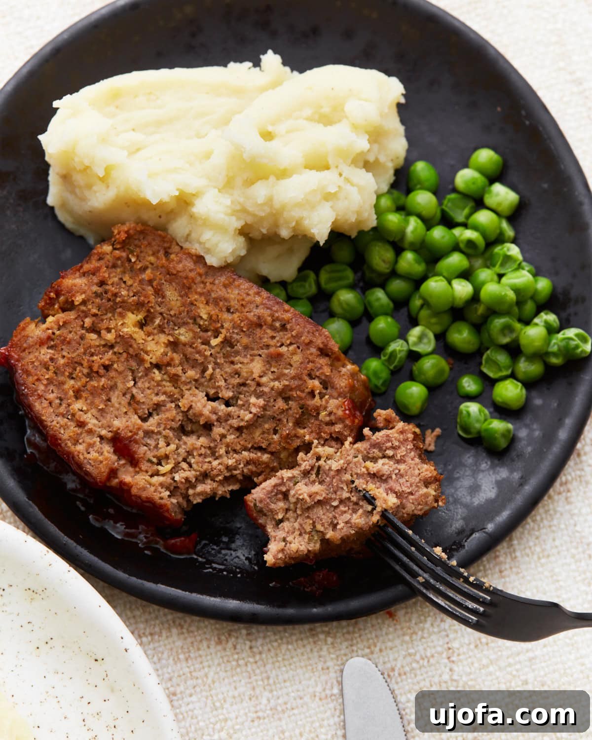 A serving of gluten-free meatloaf on a black plate with mashed potatoes and green peas, with a fork cutting into the meatloaf.