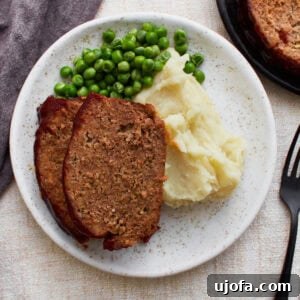 Two slices of glazed gluten-free meatloaf served with creamy mashed potatoes and green peas on a white plate, showcasing a comforting homemade meal.