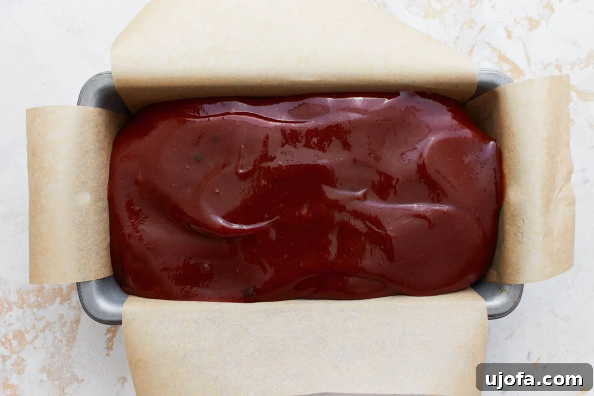 The raw gluten-free meatloaf in a parchment-lined loaf pan, topped with a layer of sweet and tangy glaze before baking.