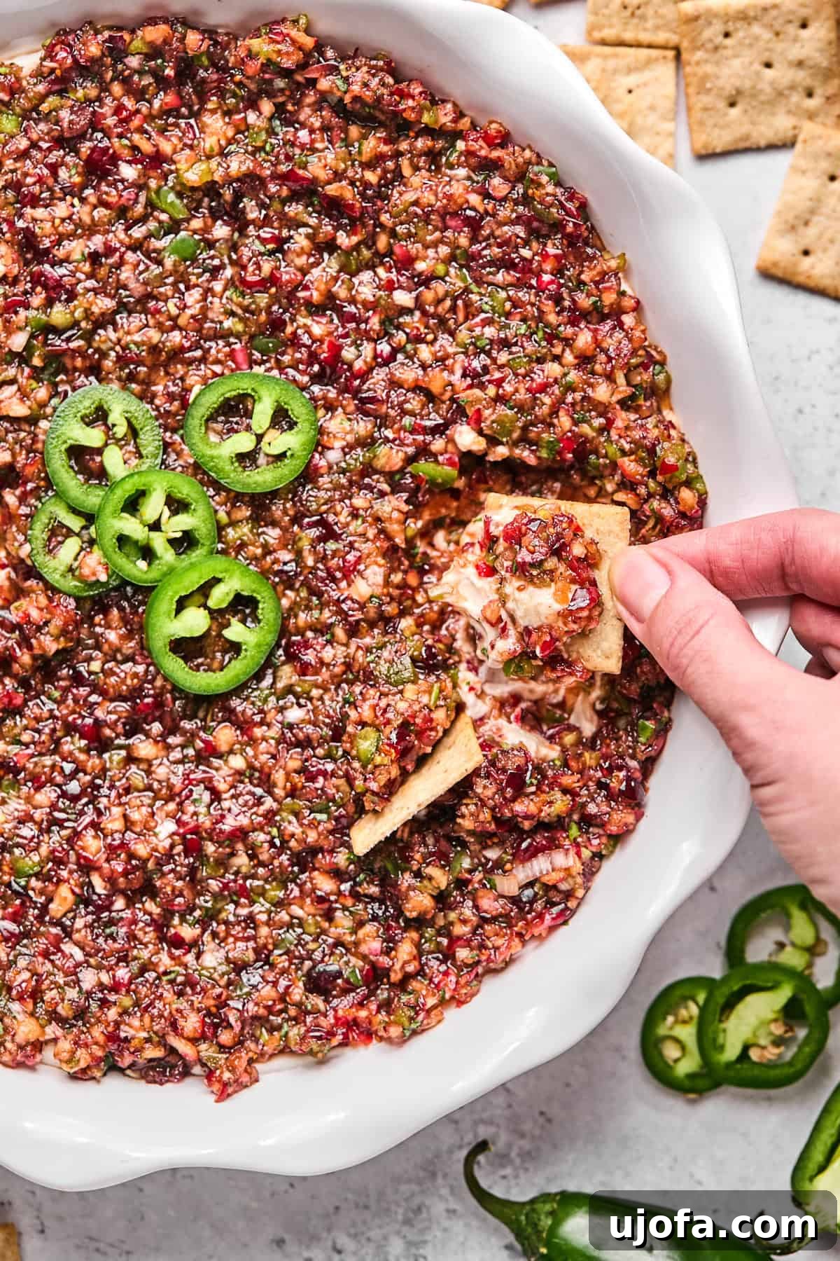 Effortless Dairy Free Cranberry Jalapeno Dip 2 cranberry jalapeno cream cheese dip in a white pie dish with a hand dipping a cracker in