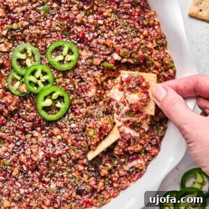 cranberry jalapeno cream cheese dip in a white pie dish with a hand dipping a cracker in