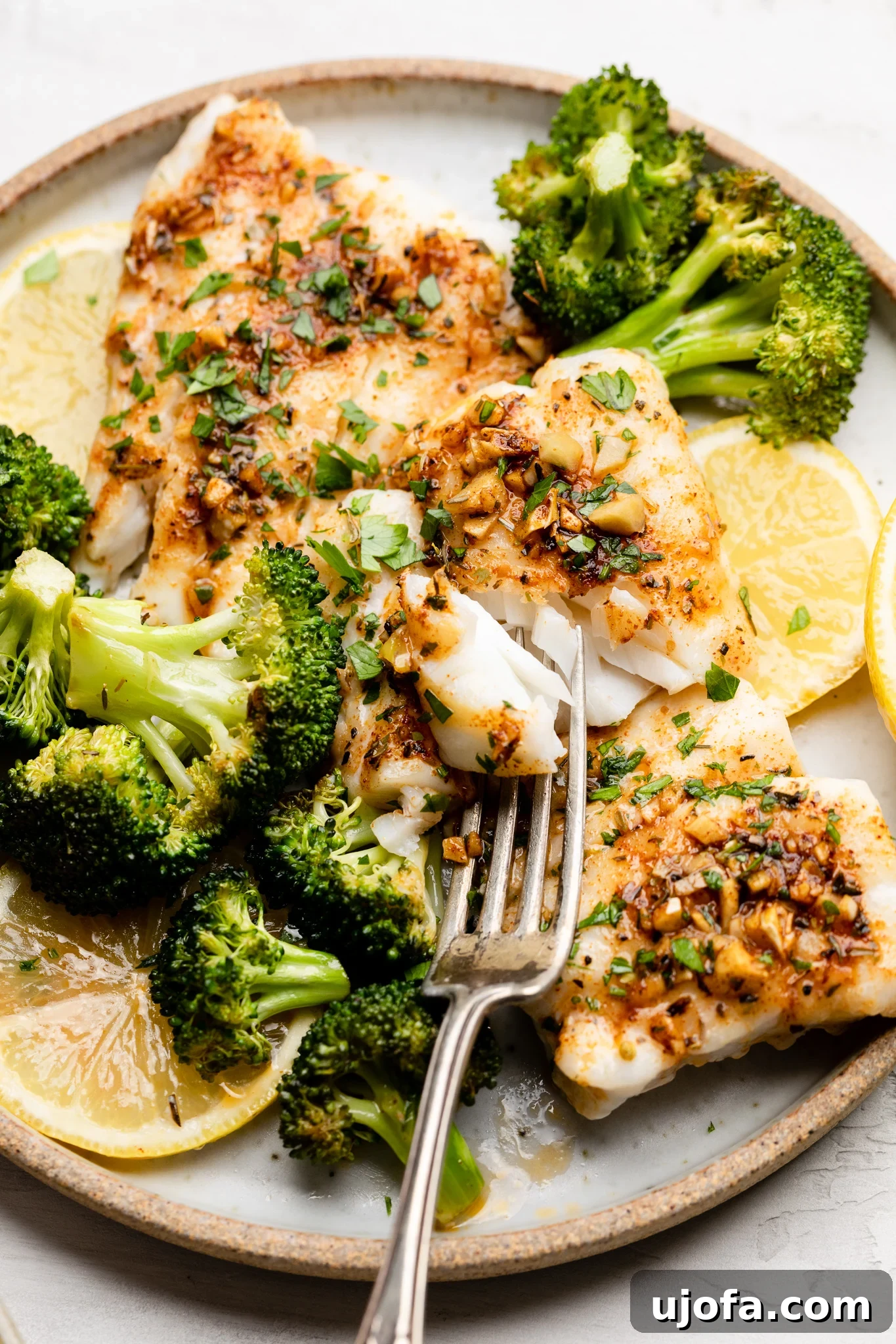 Cajun cod with broccoli garnished with fresh parsley and sliced lemons on a white plate with a silver fork, ready to eat