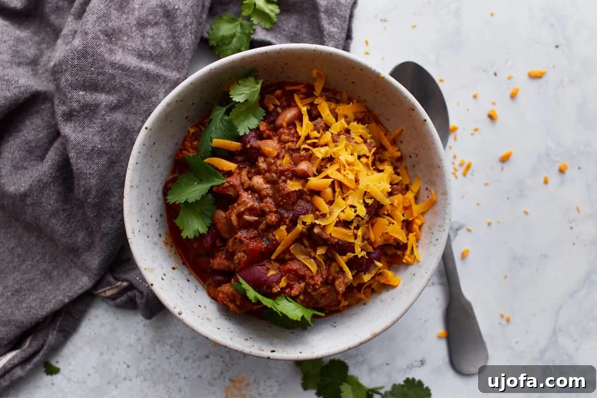 A white bowl filled with gluten-free chili, topped with parsley and shredded orange cheese. The bowl is resting on a dark grey towel with a spoon resting on the light counter beside it.