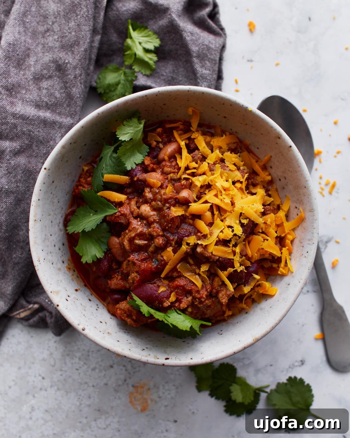 A white bowl filled with gluten-free chili, topped with parsley and shredded orange cheese. The bowl is resting on a dark grey towel with a spoon resting on the light counter beside it.