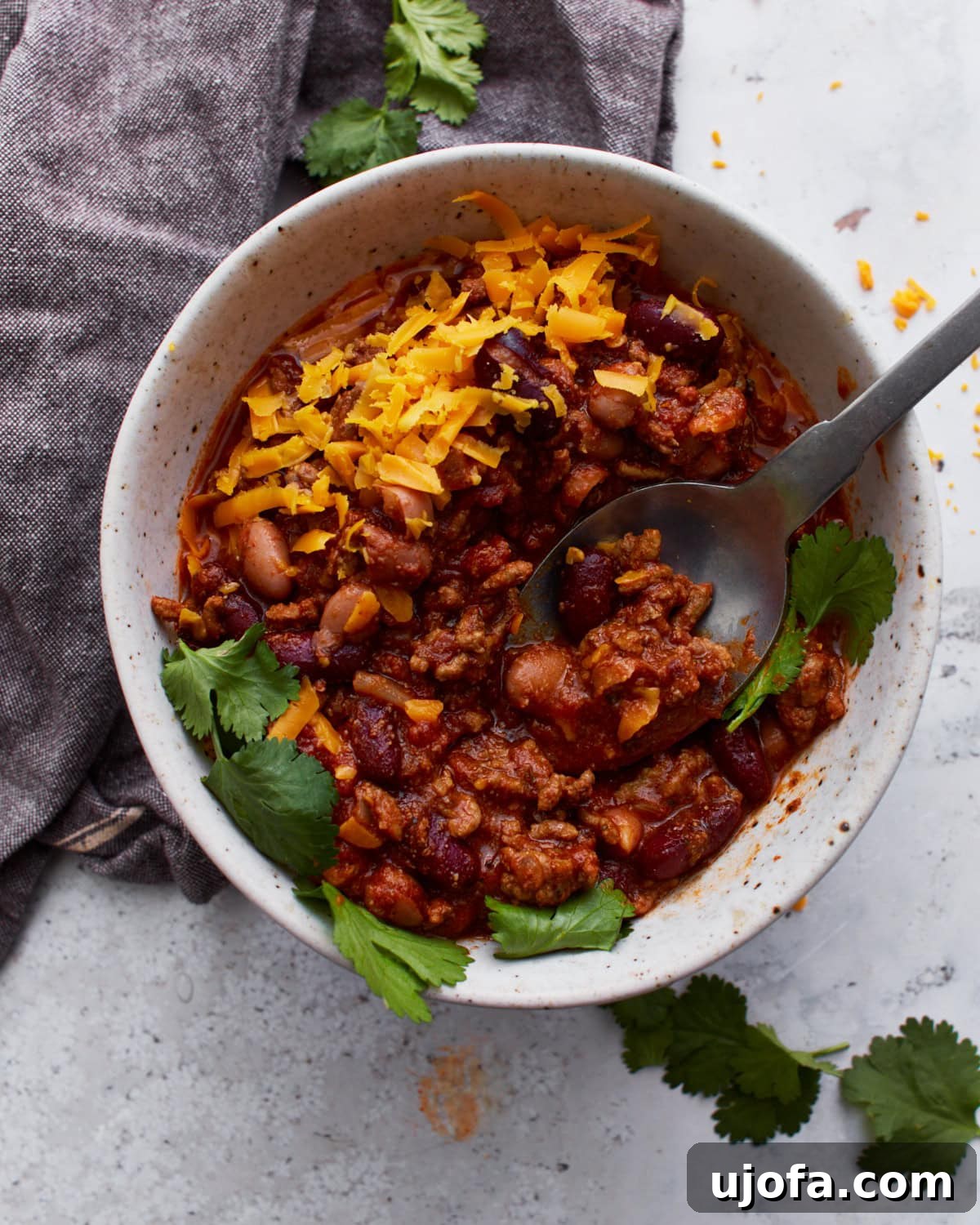 A black spoon digging into a white bowl filled with gluten-free chili, topped with parsley and shredded orange cheese.