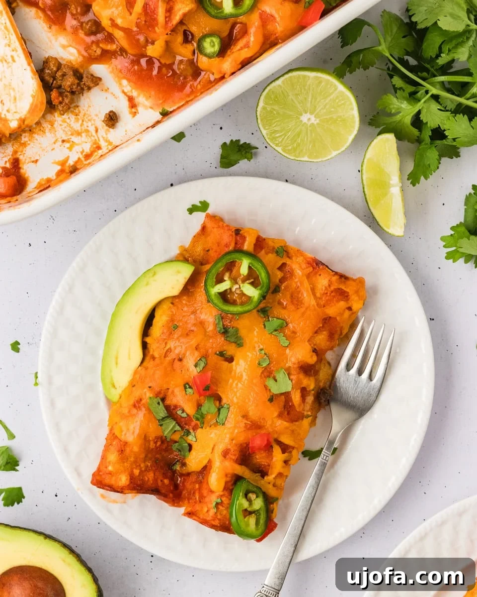 A serving of cheesy gluten-free beef and cheese enchiladas on a white plate, garnished with vibrant jalapeño slices, creamy avocado, and fresh chopped cilantro. A baking dish filled with more enchiladas and bright lime wedges are visible in the soft background.