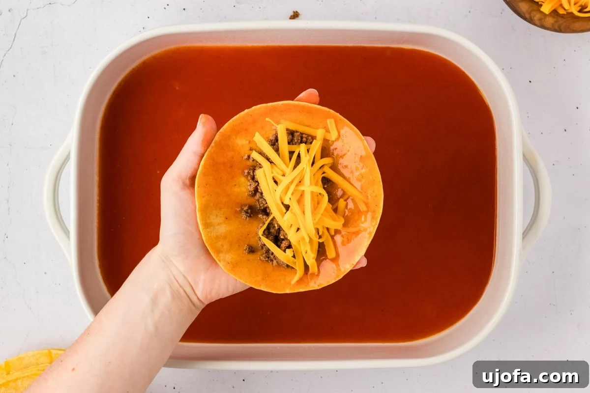A hand expertly holds a sauce-coated tortilla, which is now filled with savory seasoned beef and a sprinkle of shredded cheddar cheese, poised above the prepared baking dish of enchilada sauce.