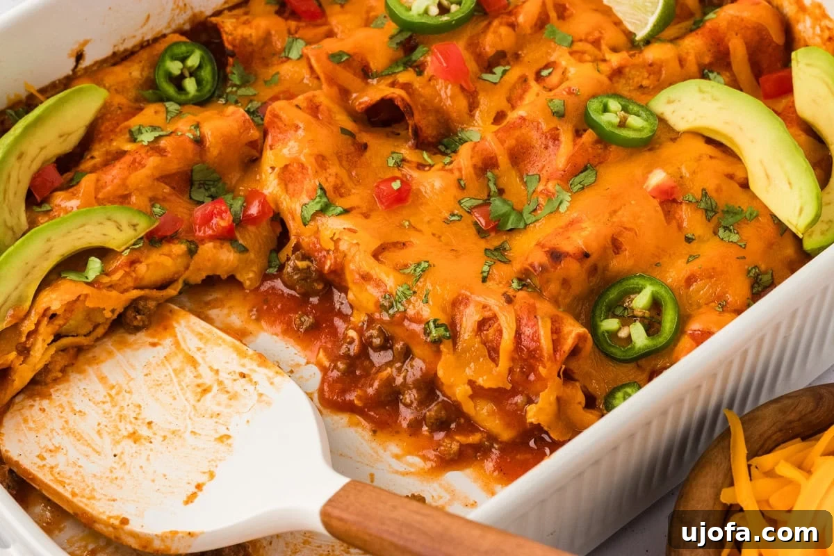 A close-up view of a baked dish of gluten-free beef and cheese enchiladas, generously topped with luscious melted cheddar, bright jalapeño slices, creamy avocado slices, vibrant diced tomatoes, and freshly chopped cilantro. A serving spatula rests invitingly in the pan, ready to dish out.