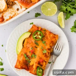A serving of cheesy gluten-free beef and cheese enchiladas on a white plate, topped with jalapeño slices, avocado, and chopped cilantro, with a baking dish and lime wedges in the background.