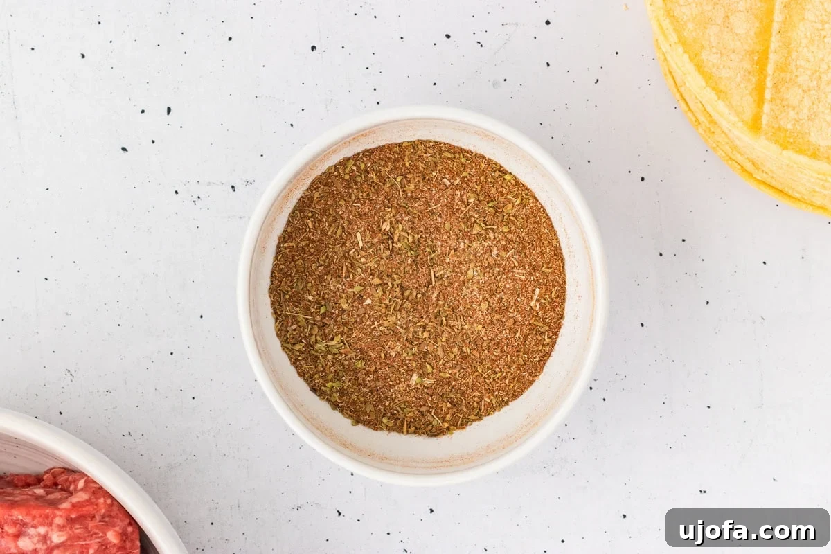 A small bowl of homemade taco seasoning mix sits on a counter, surrounded by raw ground beef and a stack of corn tortillas, highlighting the fresh components of the meal.