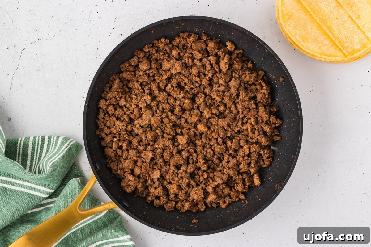 A close-up shot of a skillet filled with cooked, perfectly seasoned ground beef, awaiting assembly. A stack of golden corn tortillas and a green striped kitchen towel rest nearby.