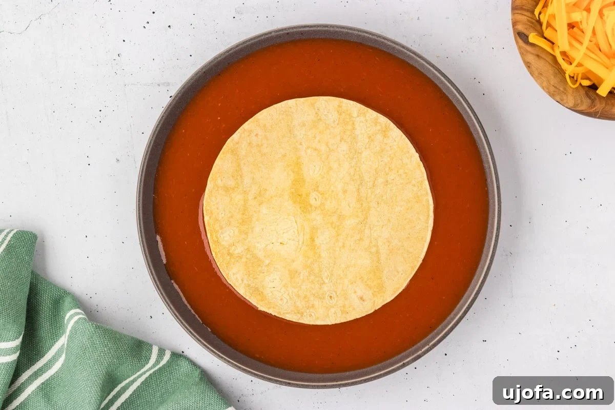 A corn tortilla is gracefully soaking in a shallow bowl of rich red enchilada sauce, preparing it for filling. A wooden bowl of shredded cheddar cheese waits in the background.