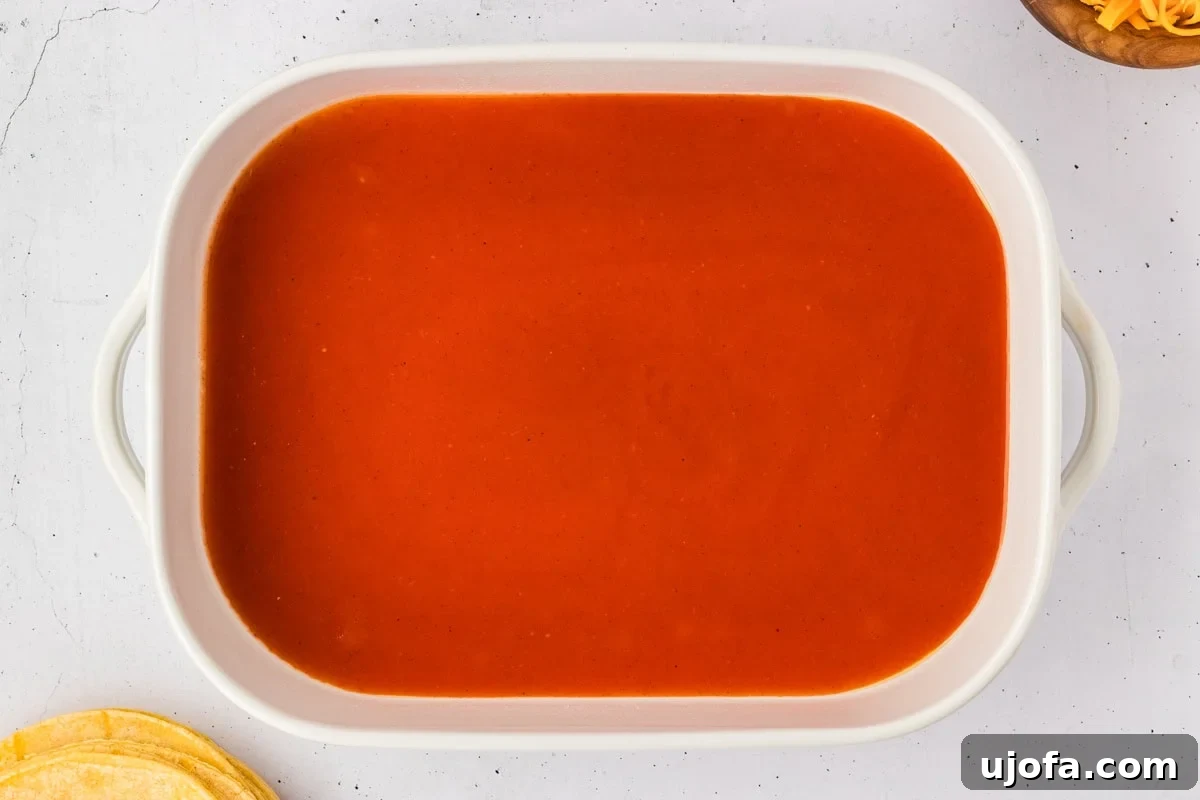 A pristine 9x13 white baking dish, its bottom evenly coated with a vibrant layer of red enchilada sauce, perfectly prepped for the enchilada assembly.