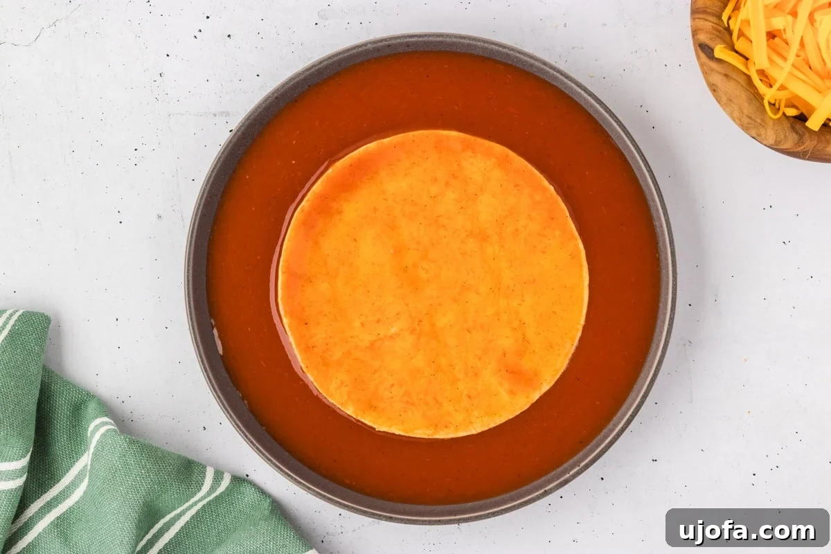 A single corn tortilla, fully saturated with rich red enchilada sauce, waits in a shallow dish, next to a bowl of freshly shredded cheddar cheese, ready for the next step in the assembly process.