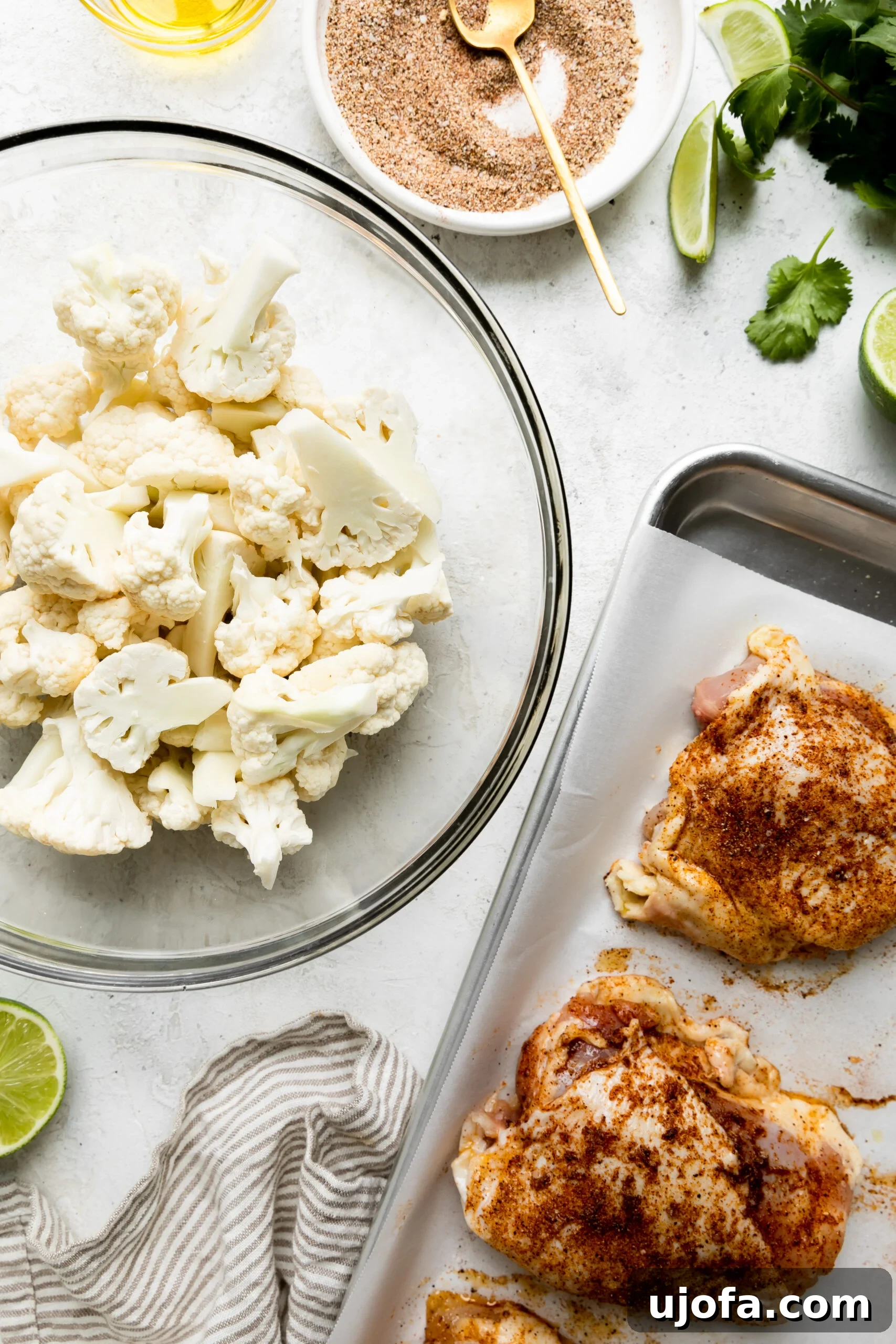A close-up photo of raw cauliflower in a glass bowl alongside chicken thighs arranged on a silver baking pan, with a small bowl of vibrant cilantro lime sauce ready for drizzling.