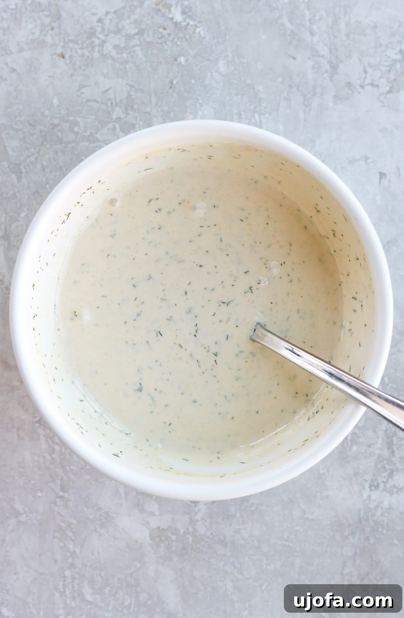 A smooth, creamy mayonnaise-based sauce sits in a pristine white bowl, accompanied by a gleaming silver spoon, awaiting its role in the pasta salad.