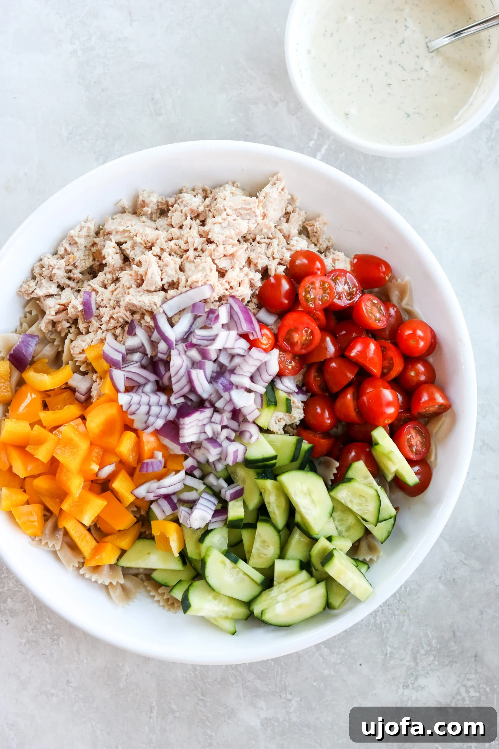 A mix of fresh ingredients arranged in a white bowl, positioned next to another white bowl containing a creamy mayonnaise sauce, highlighting the components of the healthy pasta salad.