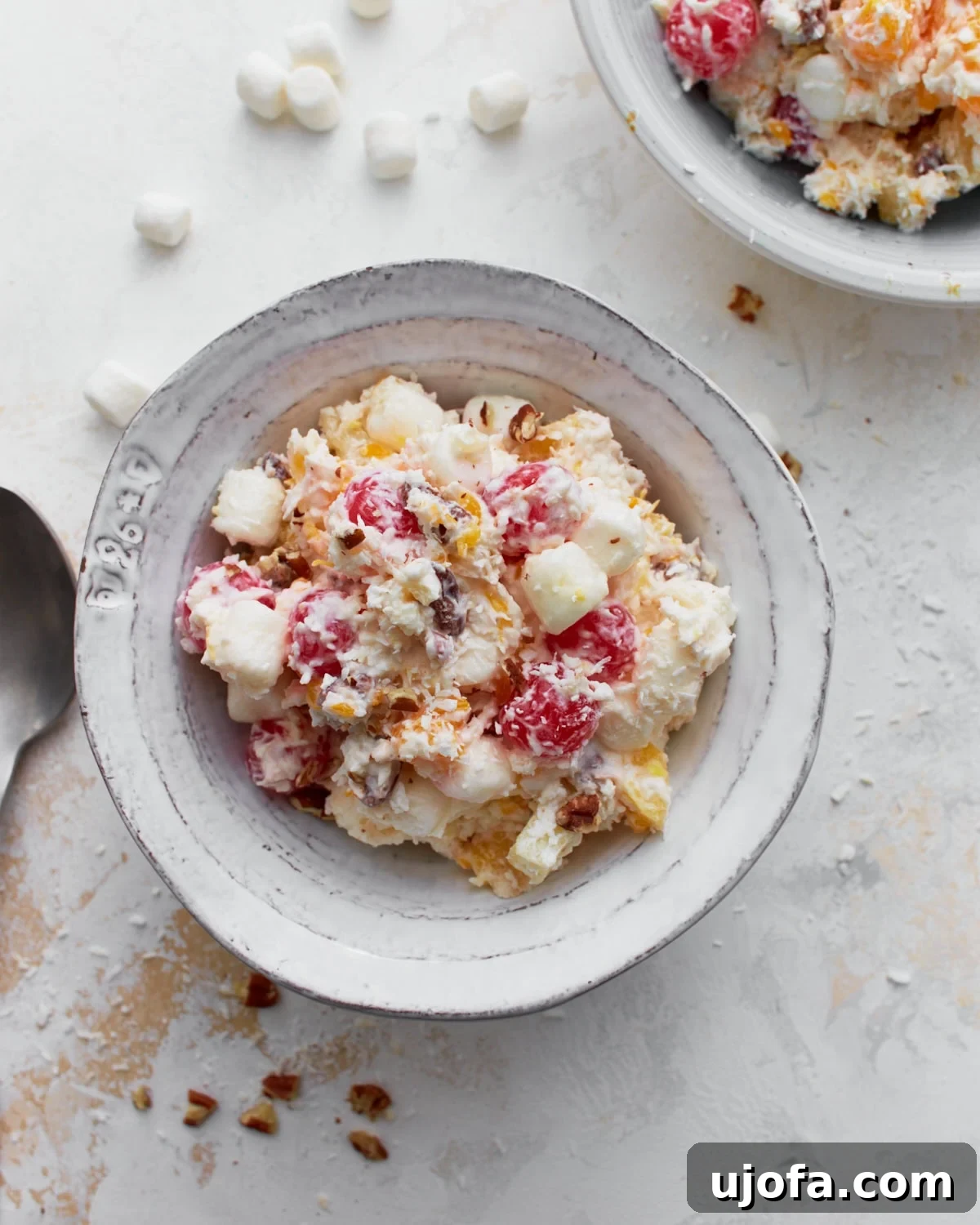 Divine Fruit Ambrosia 2 A vibrant bowl of Ambrosia Salad, showcasing a rich blend of creamy whipped topping, juicy pineapple chunks, bright mandarin oranges, ruby-red maraschino cherries, soft mini marshmallows, delicate shredded coconut, and crunchy chopped nuts. Ready to be served.