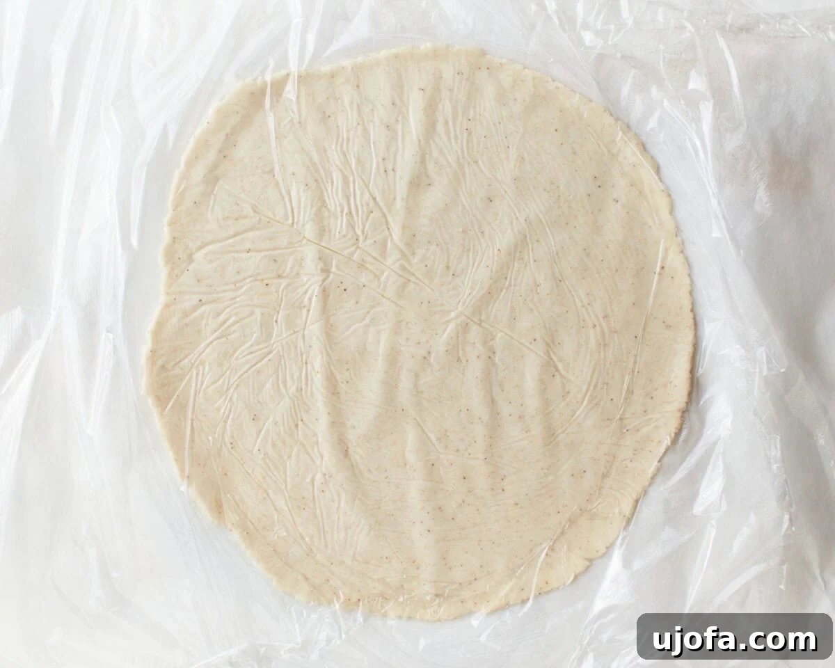 A gluten-free tortilla dough being rolled thinly between two pieces of plastic wrap with a rolling pin, ensuring even thickness and preventing sticking.