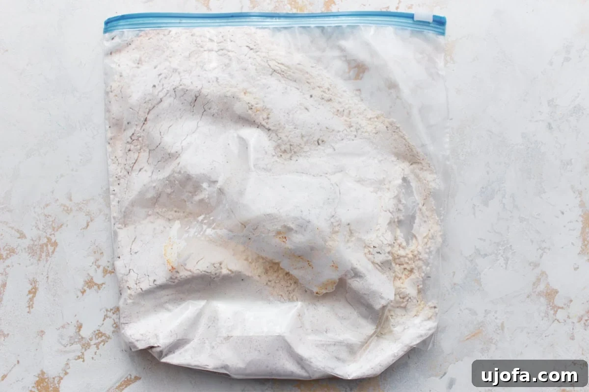 Chicken thighs and drumsticks covered in gluten-free flour, cornstarch, and spice mixture in a large plastic bag laid flat on a light surface.