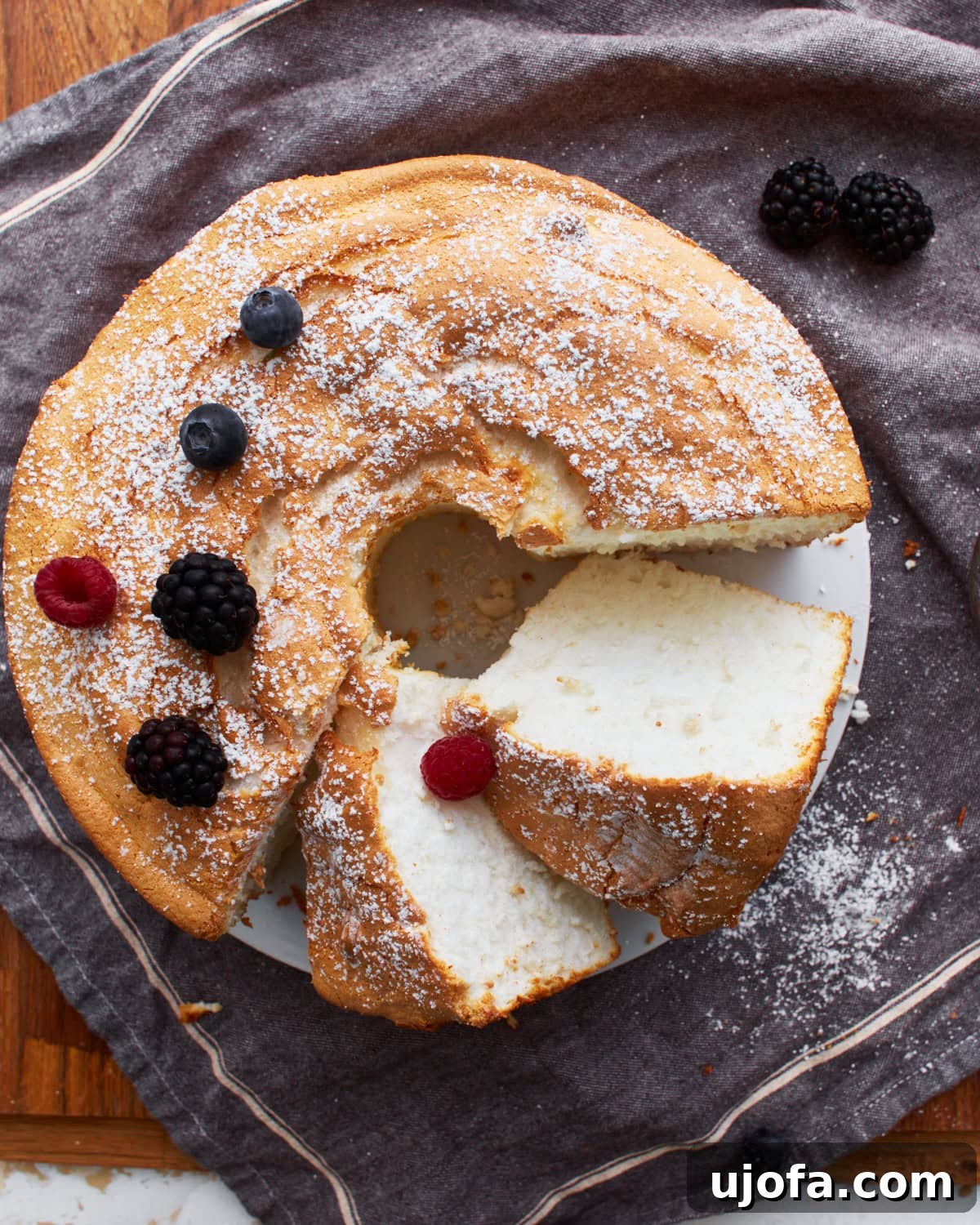 Sliced gluten-free angel food cake on a grey towel, dusted with powdered sugar and garnished with fresh berries.