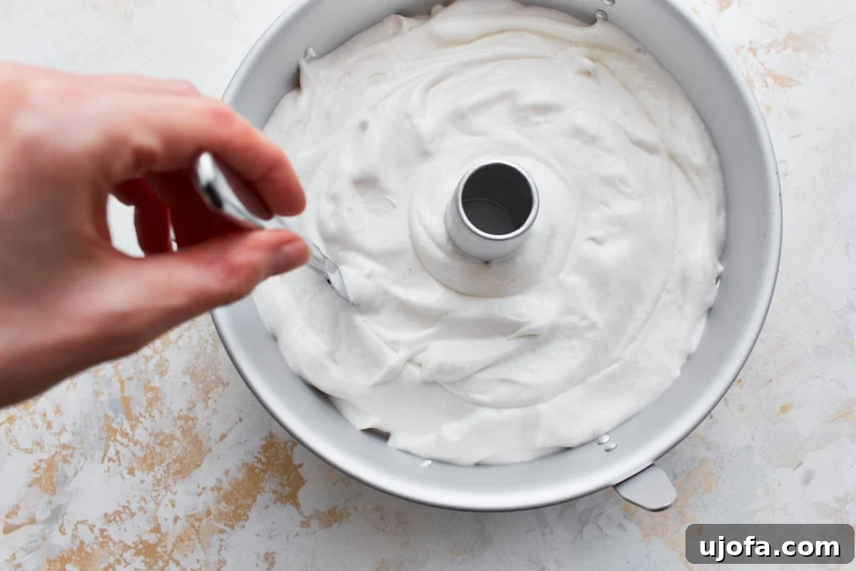 A hand using a knife to gently cut through gluten-free angel food cake batter in a tin tube pan to release air bubbles.