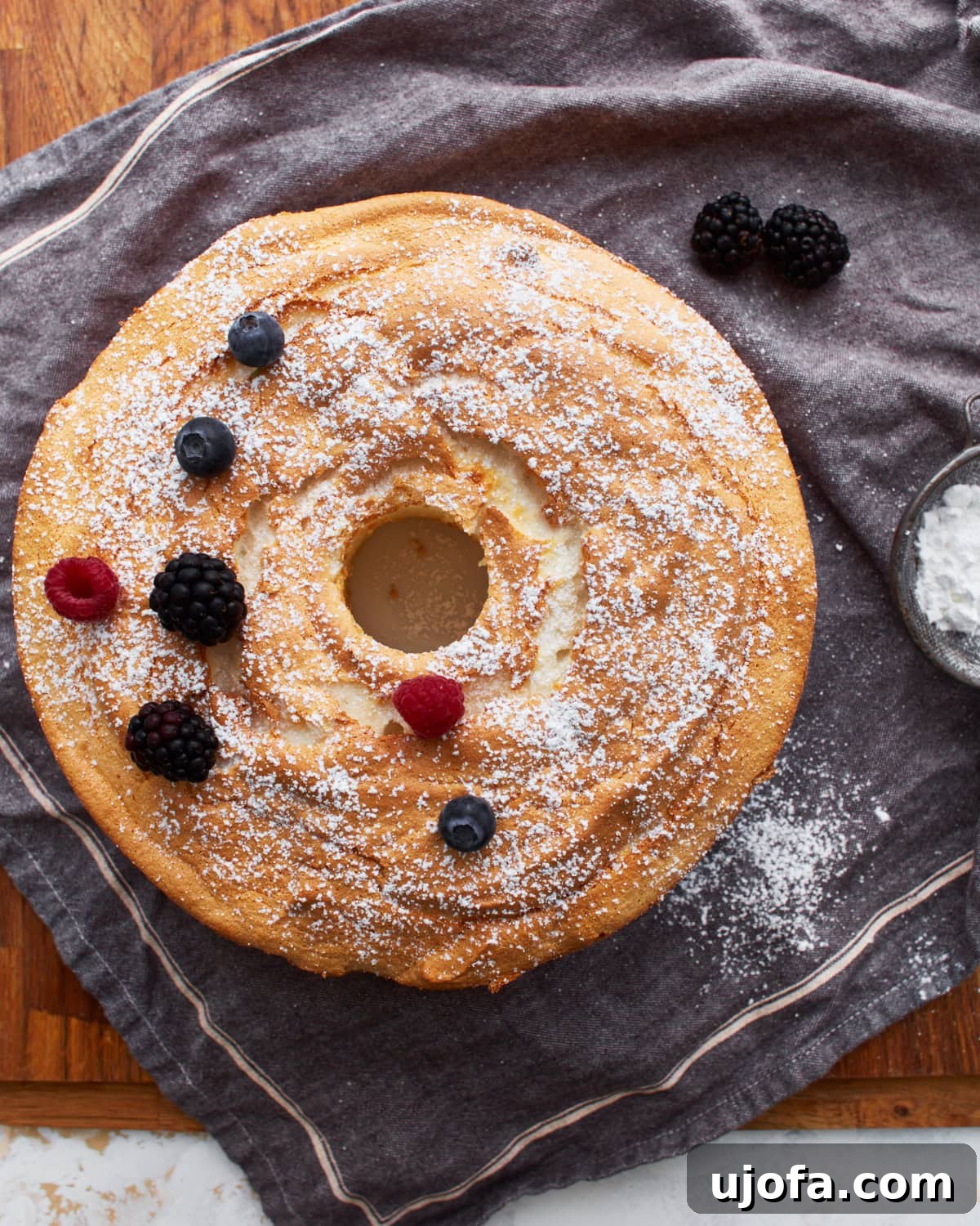 A whole gluten-free angel food cake on a grey towel, sprinkled with powdered sugar and adorned with blackberries, raspberries, and blueberries.