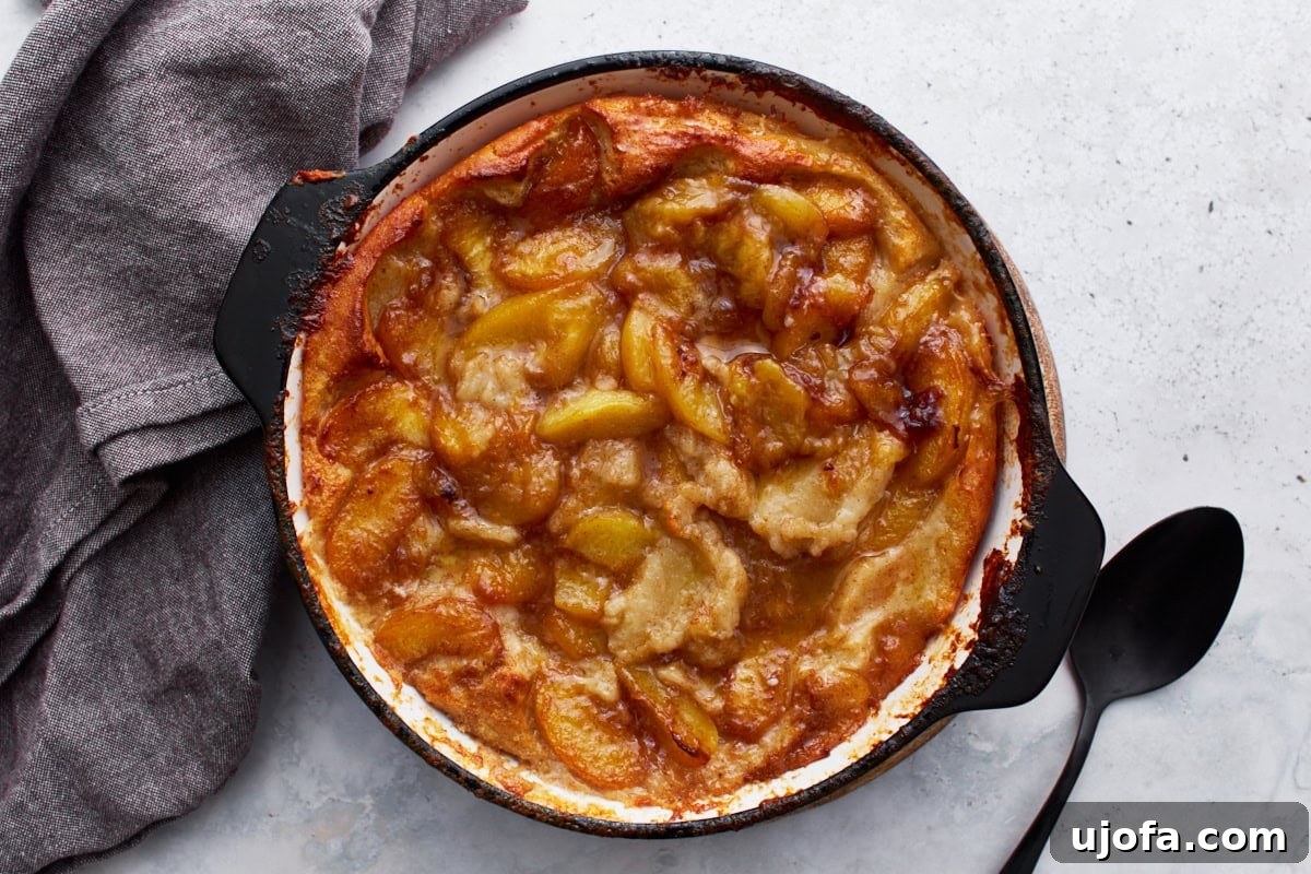 Heavenly Gluten-Free Peach Cobbler 11 A baked peach cobbler in a black skillet, perfectly golden brown, resting on a wooden surface beside a gray napkin and a black serving spoon.