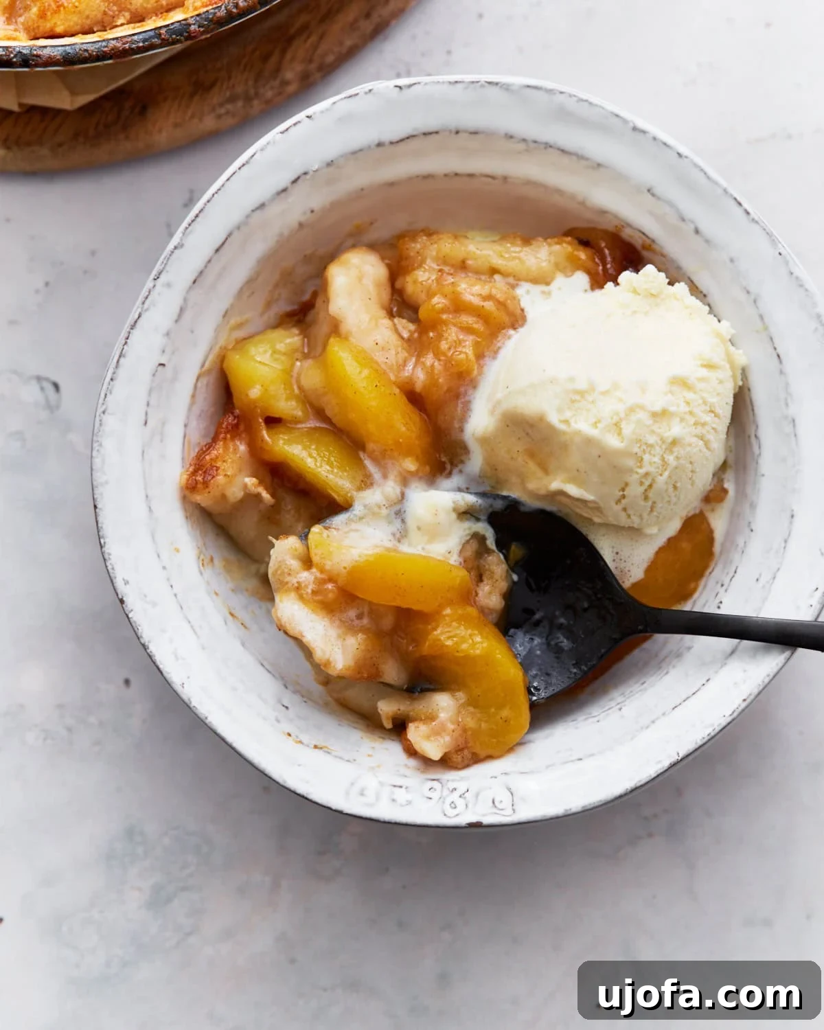 Heavenly Gluten-Free Peach Cobbler 13 A close-up of a spoonful of gluten-free peach cobbler being lifted from a white bowl, revealing tender peaches, a golden crust, and slowly melting vanilla ice cream.