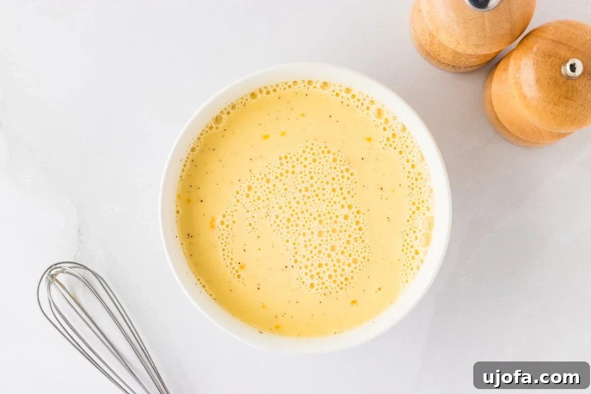 Ultimate Gluten-Free Quiche 6 A smooth, light yellow mixture of whisked eggs and milk in a glass bowl, with a whisk resting on the side and wooden salt and pepper shakers in the background.