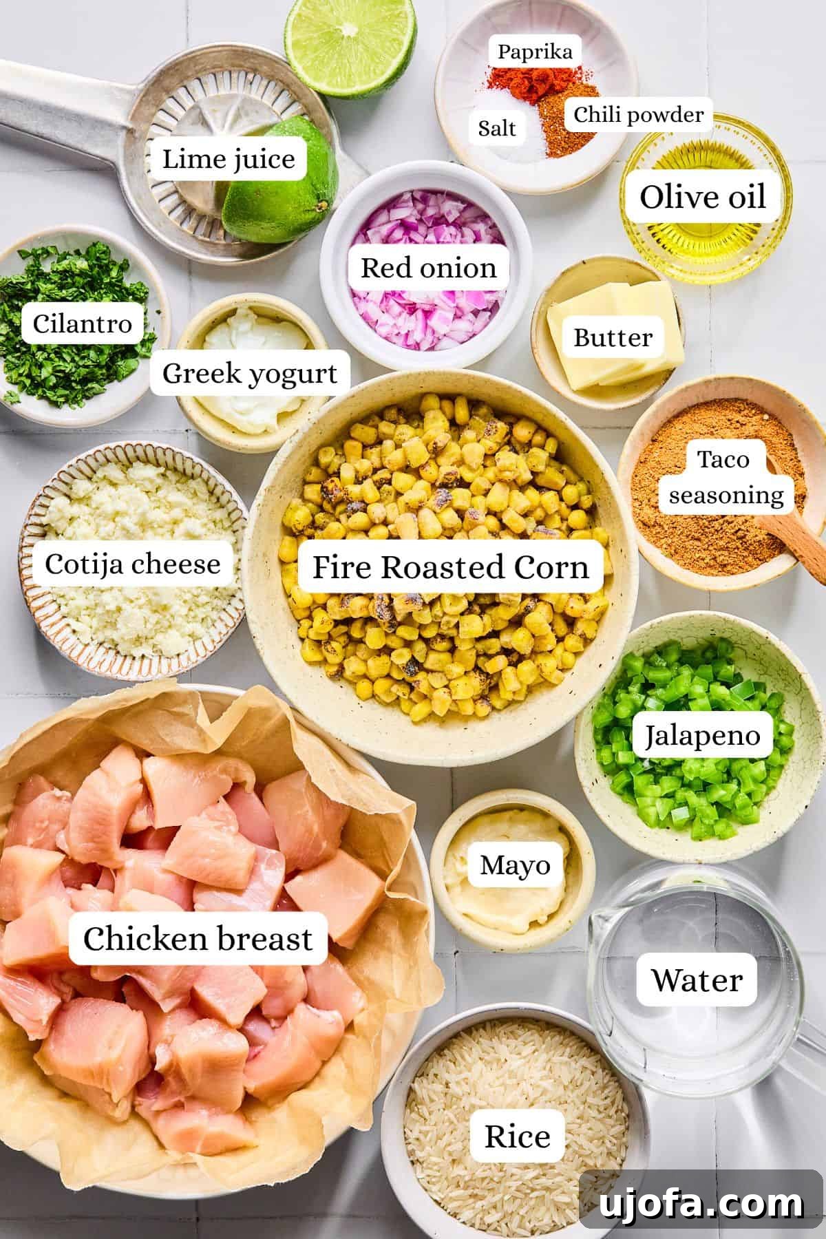 Various fresh ingredients for street corn chicken rice bowls, including corn, red onion, jalapeño, cilantro, lime, and chicken breast, neatly arranged in small white bowls on a pristine white countertop.