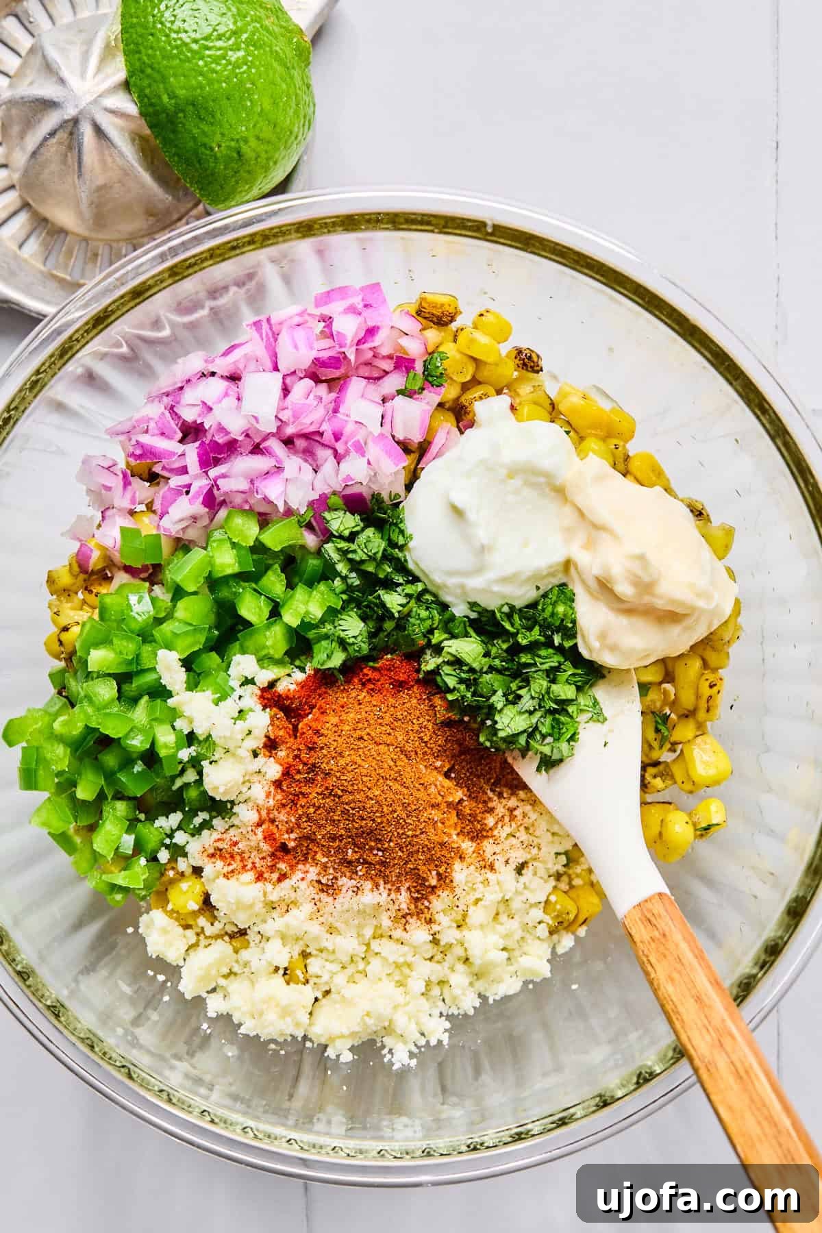 Creamy street corn mixture being stirred in a glass bowl with diced red onion, jalapeño, cilantro, mayonnaise, Greek yogurt, lime juice, various spices, and crumbled cotija cheese, ready for assembly.
