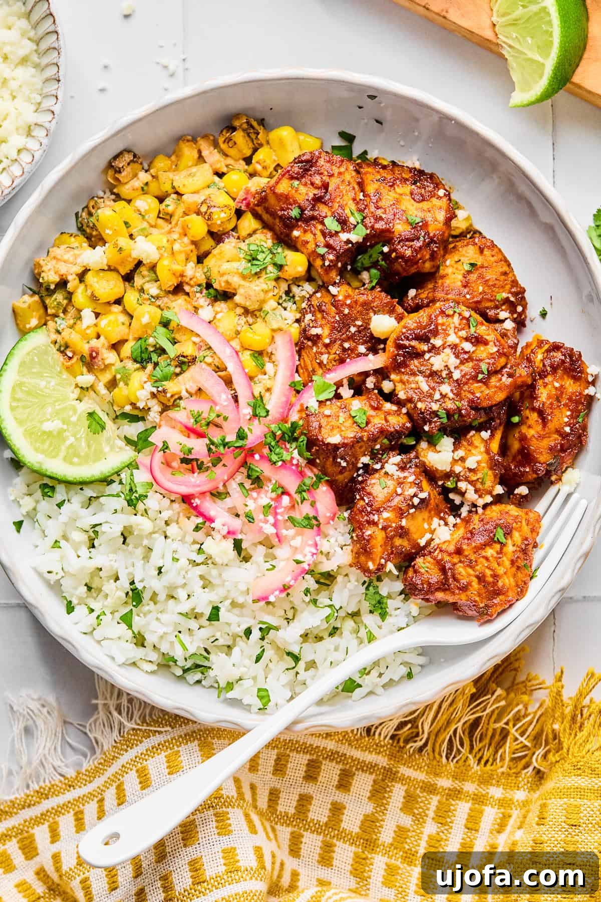A beautifully assembled street corn chicken and rice bowl, garnished with cilantro and a lime wedge, with a fork ready to dig in, placed on a yellow napkin.
