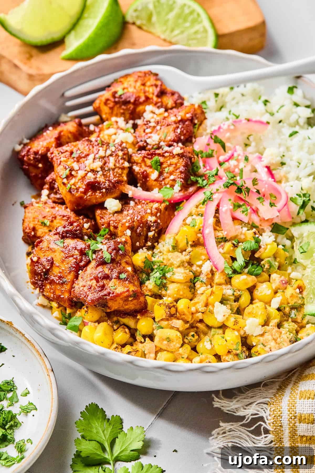 Close-up view of a street corn chicken rice bowl, with a fork lifting a piece of perfectly cooked taco-seasoned chicken, beautifully garnished with fresh cilantro and a lime wedge.