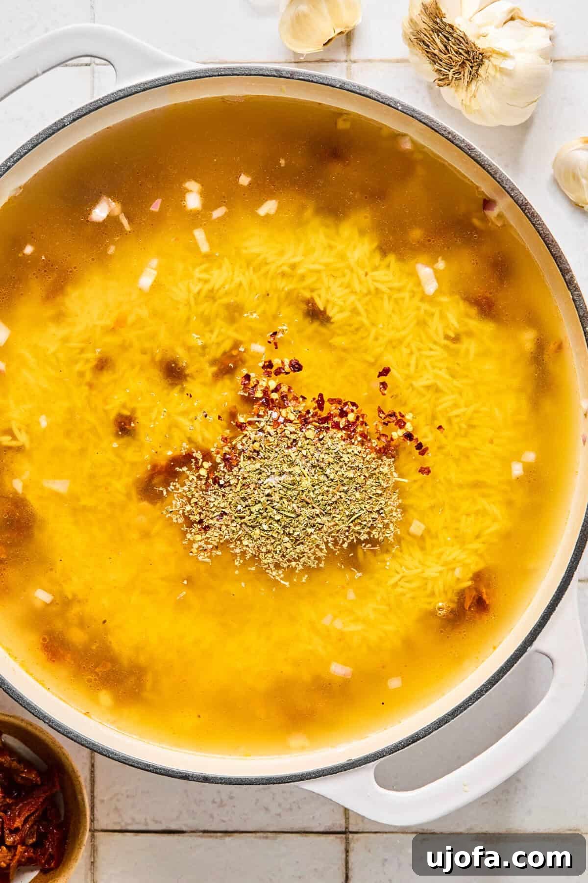 Gluten-free orzo simmering in chicken broth with Italian seasoning and red pepper in a skillet.