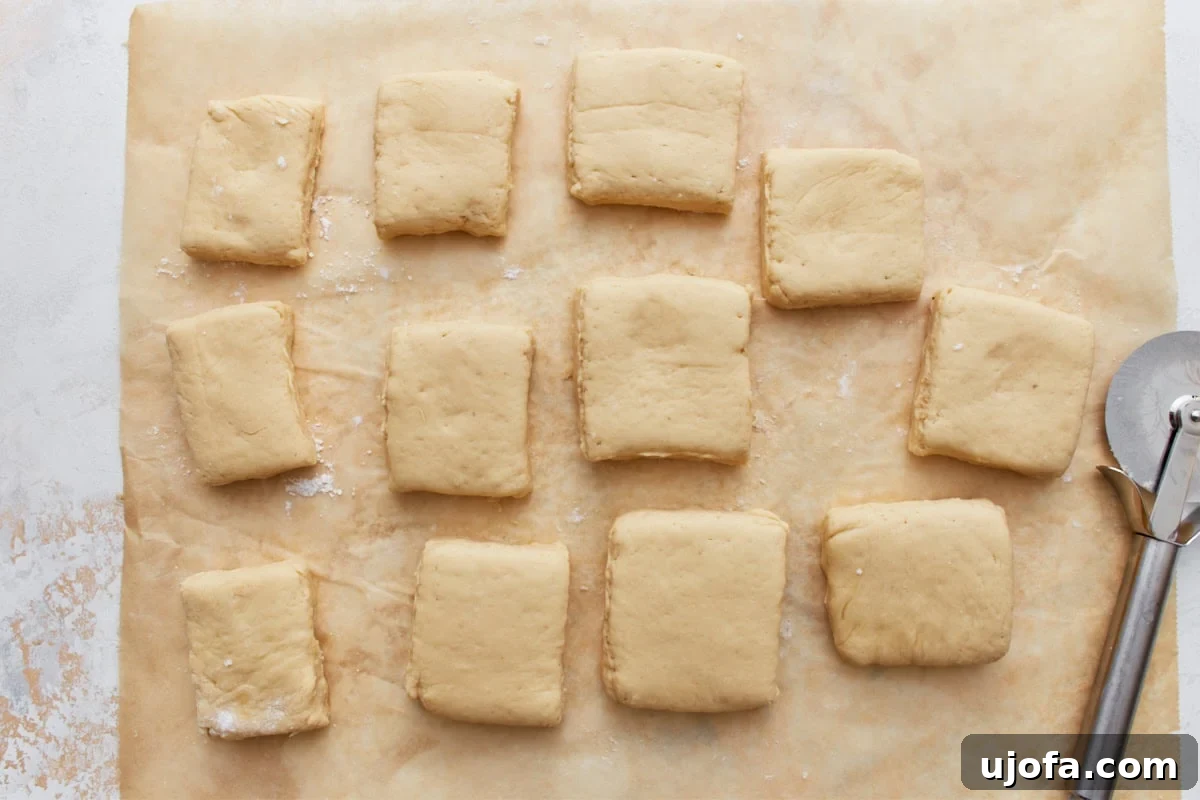 Pillowy Gluten-Free Beignets 10 Squares of gluten-free beignet dough cut and spaced on parchment paper with a pizza cutter nearby.