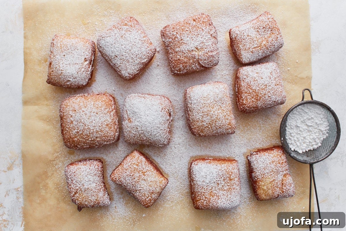 Pillowy Gluten-Free Beignets 13 Freshly fried gluten-free beignets dusted with powdered sugar, arranged on parchment paper with a mesh sifter on the side.