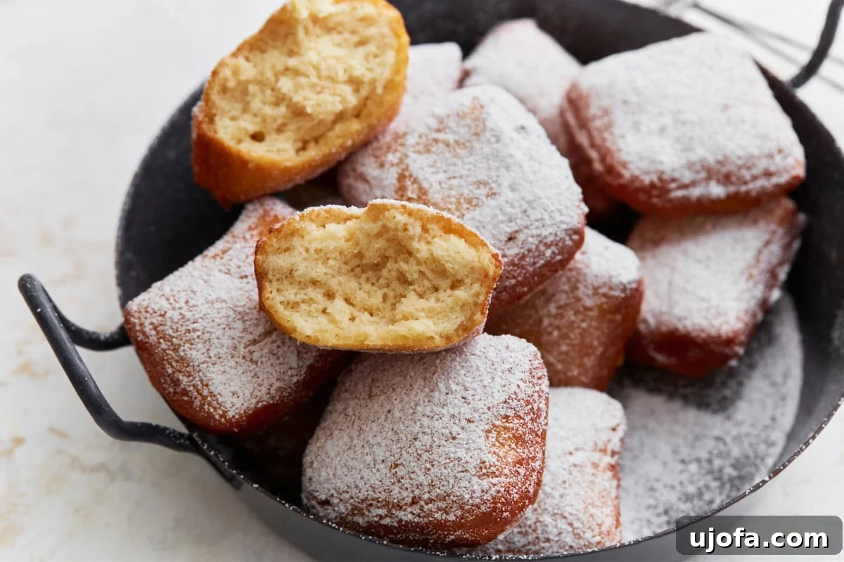 Pillowy Gluten-Free Beignets 14 A close-up of a halved gluten-free beignet showing its fluffy interior, stacked on top of more powdered sugar–coated beignets in a black serving dish.