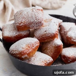 Pillowy Gluten-Free Beignets 3 A pile of golden brown gluten-free beignets dusted with powdered sugar, served in a black pan.