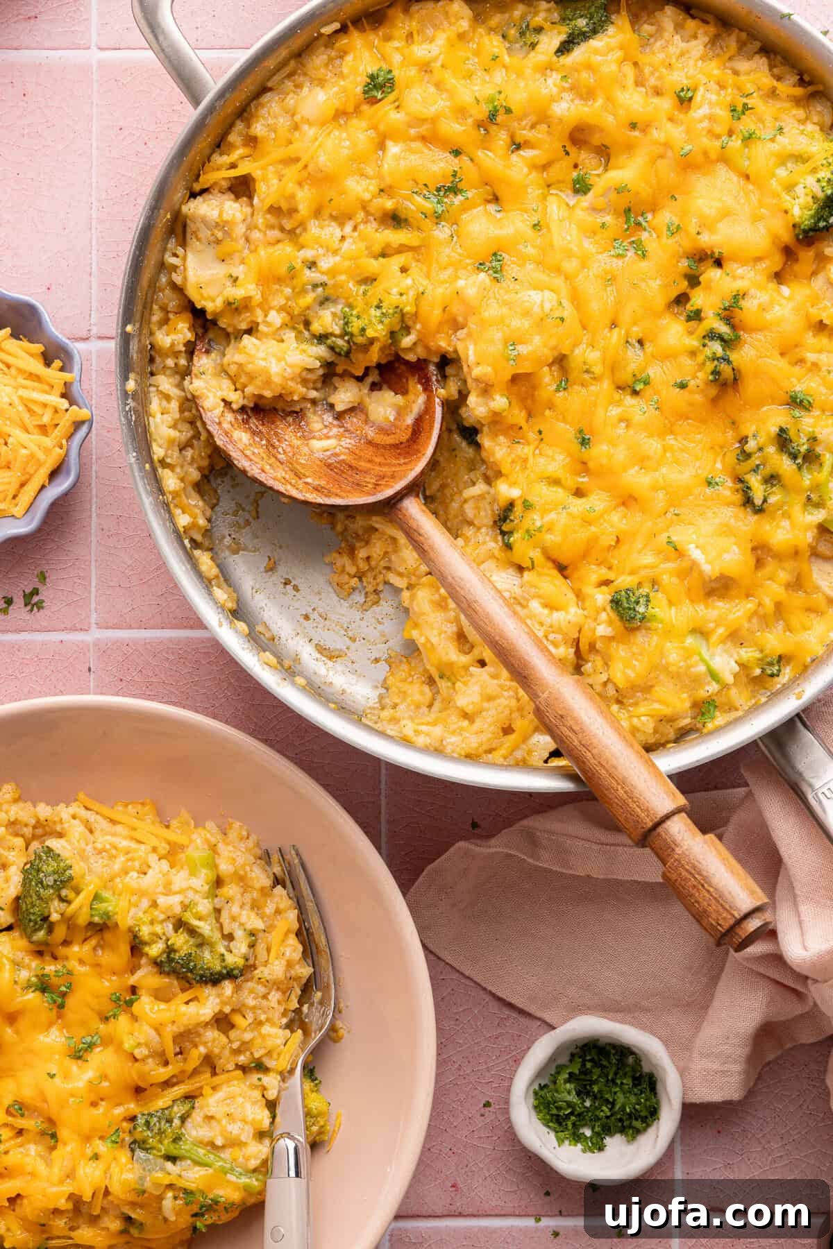 Ultimate Chicken Broccoli Rice Bake 10 A large skillet brimming with cheesy chicken broccoli rice casserole and a wooden spoon, alongside a serving on a white plate.