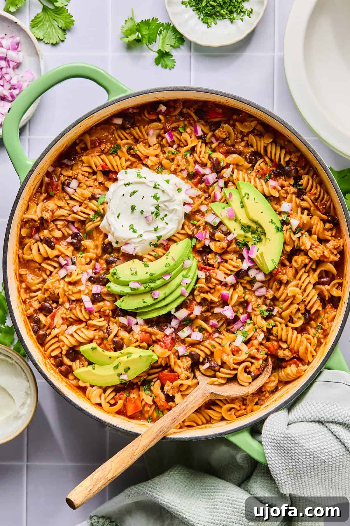 One pot taco pasta in the pot topped with sour cream, avocado, and cilantro with a wooden spoon, ready to be served.