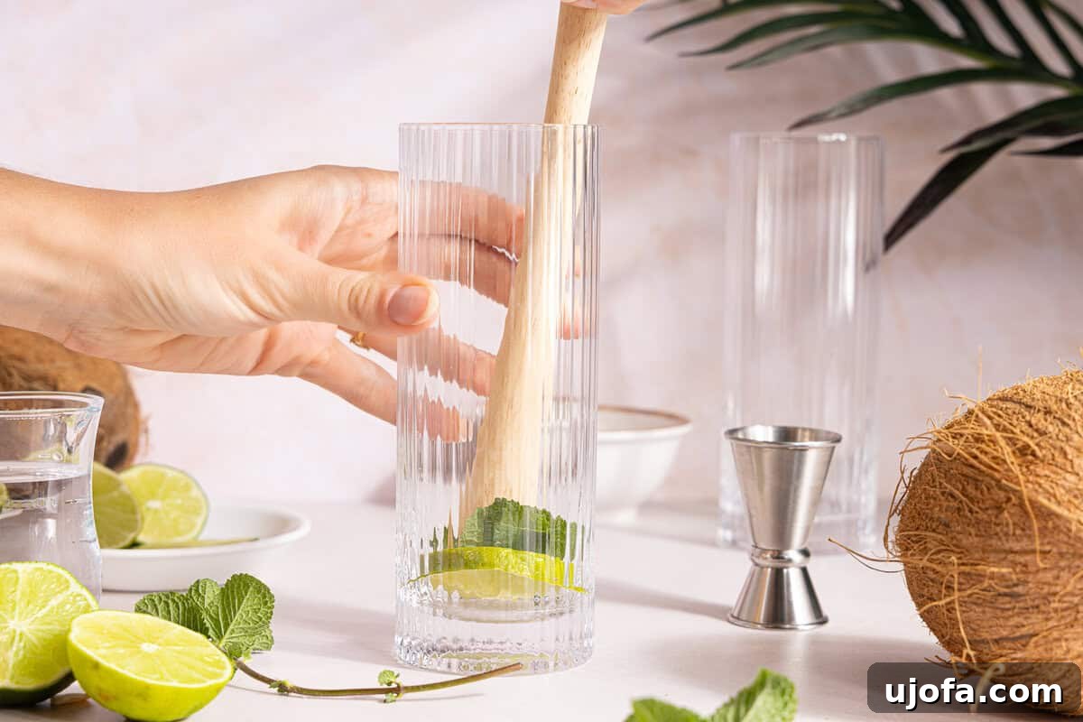 Dreamy Coconut Mojito 4 A muddler gently pressing a lime wedge and fresh mint leaves at the bottom of a tall glass, preparing the base for a mojito.