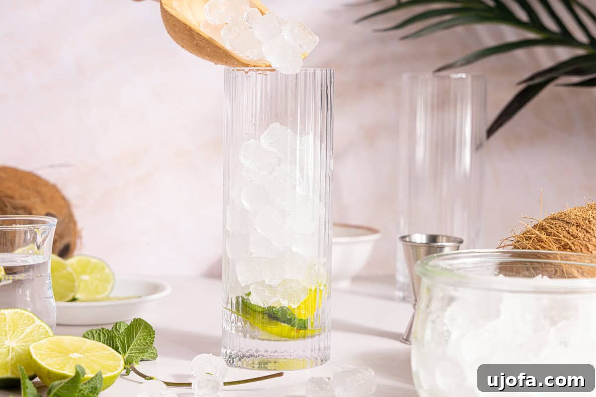 Dreamy Coconut Mojito 5 Ice cubes being poured into a tall glass already containing muddled mint and lime, filling it three-quarters of the way.