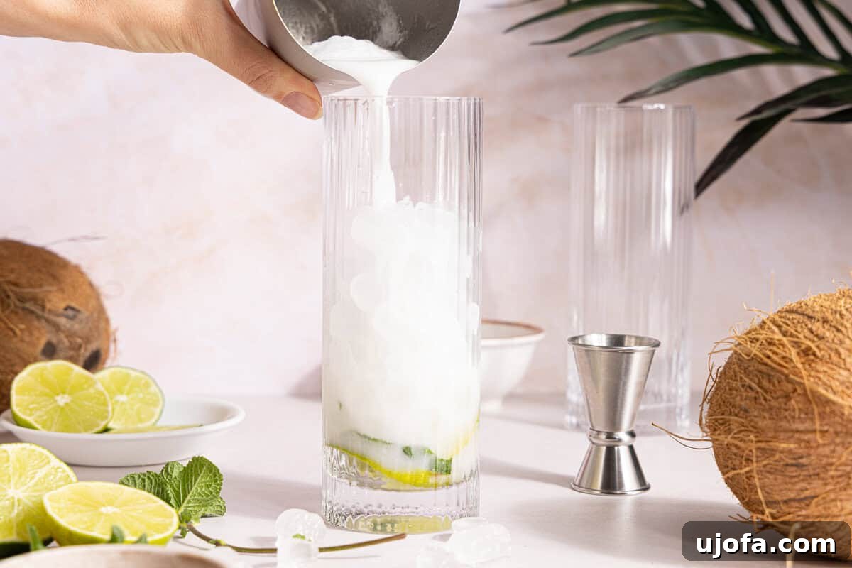 Dreamy Coconut Mojito 7 The creamy white rum and cream of coconut mixture being poured from a cocktail shaker over ice in the glass with muddled mint and lime.