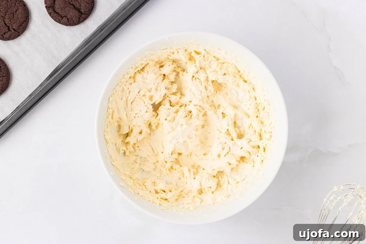 A white bowl filled with fluffy vanilla marshmallow cream filling, ready to be piped onto cookies. A baking tray with chocolate cookies and a whisk are partially visible nearby.