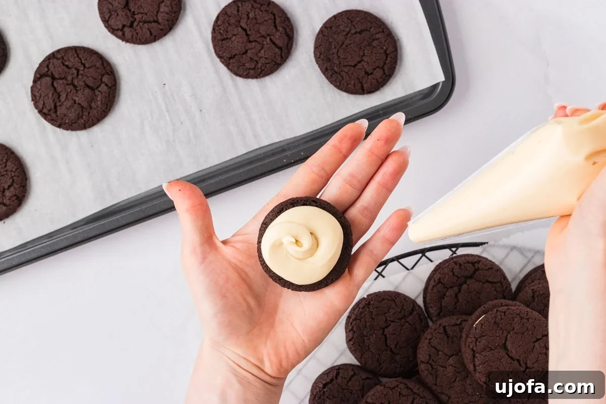 A hand holding a chocolate cookie with piped vanilla filling, using a piping bag to prepare the sandwich cookies on a cooling rack.